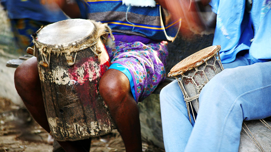 A close-up of two people in colorful clothing playing traditional African talking drums, their hands blurred with motion.