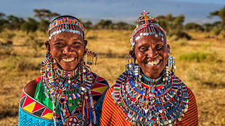 Two Maasai women in traditional colorful beaded clothing and jewelry smile together in a grassy field in Kenya.