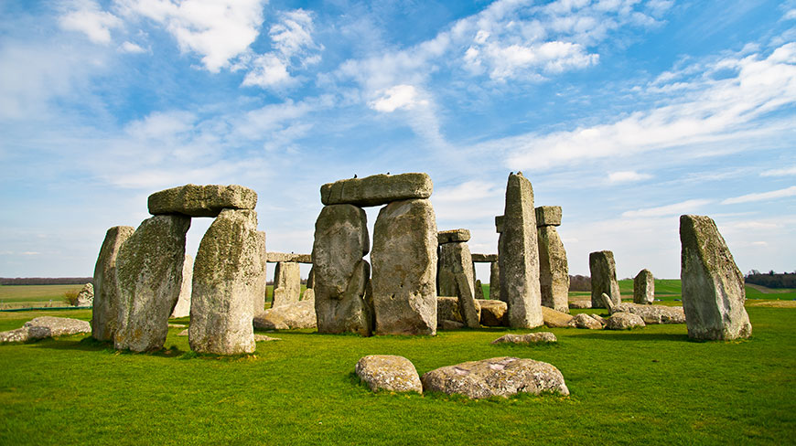 The ancient stone circle of Stonehenge stands in a green field under a partly cloudy blue sky.