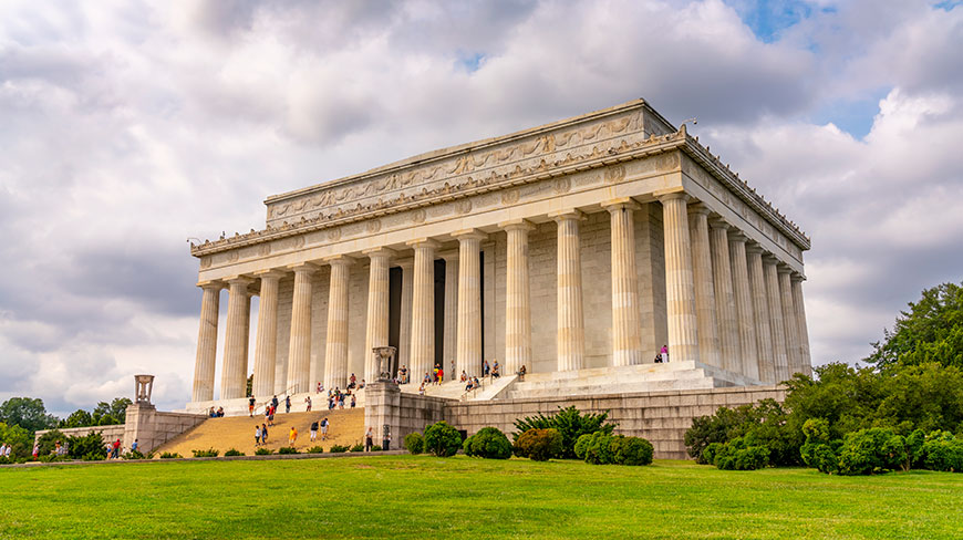 The Lincoln Memorial in Washington, D.C., is viewed from the lawn with visitors on the steps under a cloudy sky.