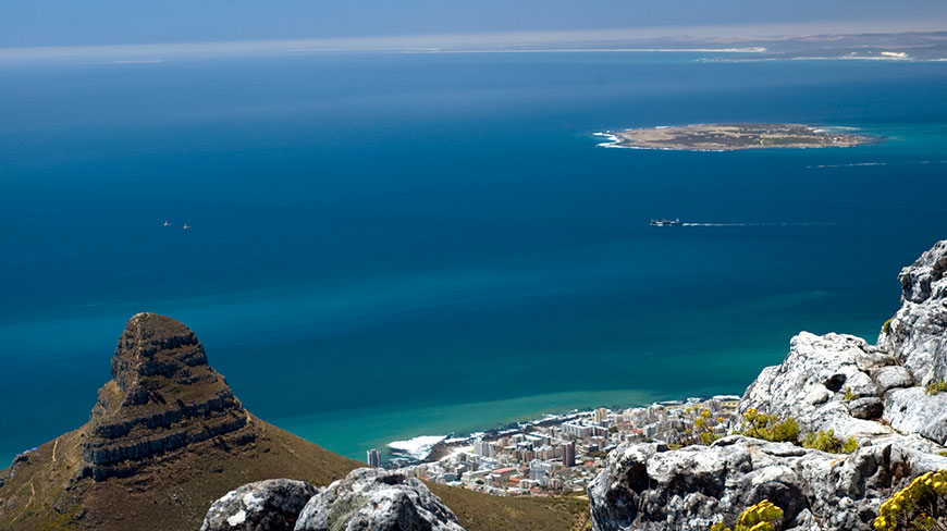 A high-angle view of Cape Town, South Africa, shows Lion's Head peak and Robben Island in the distance across the blue Atlantic Ocean.