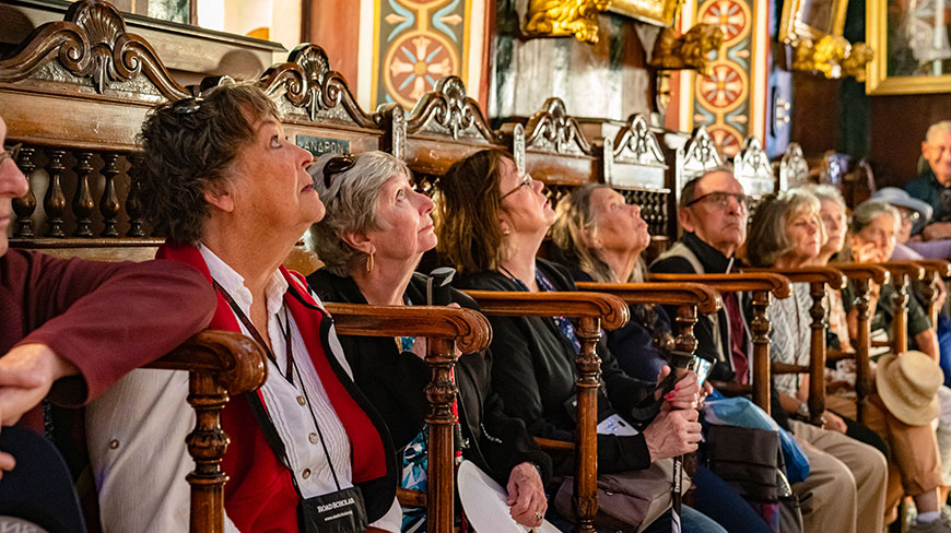 A group of travelers on a tour in Greece sit in ornate wooden pews, looking up with interest inside a historic building in Ermoupoli.