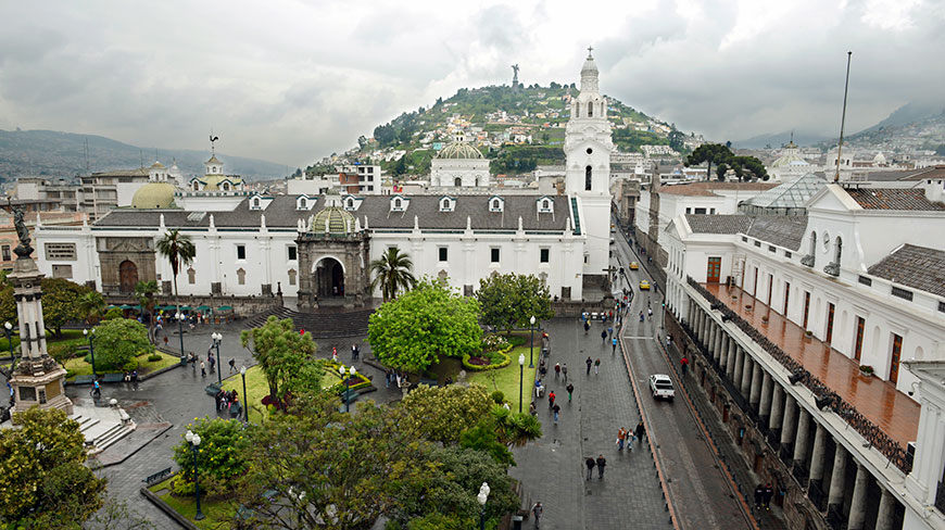 An aerial view of a bustling city square in Quito, Ecuador, with historic white buildings and a green hill in the background.
