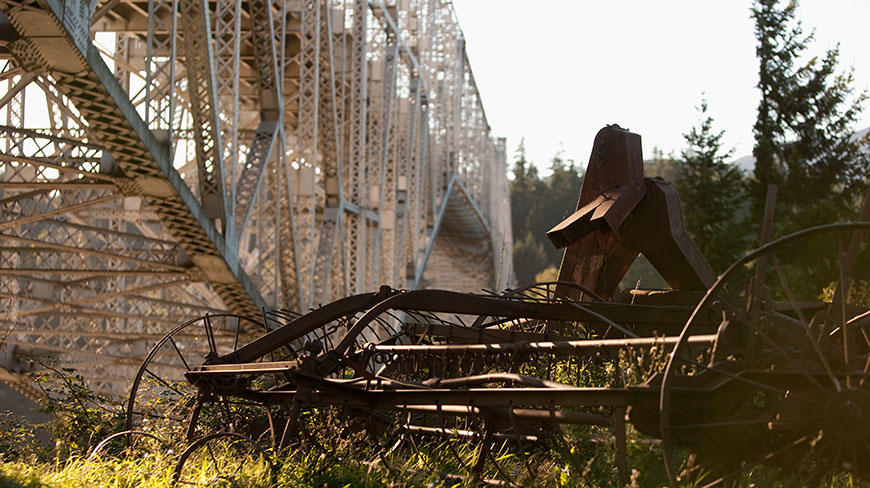 Sunlight shines on rusted antique farm equipment in a grassy field below a large steel bridge in Cascade Locks, Oregon.