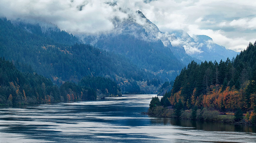 A wide river flows through the forested Columbia River Gorge in Oregon, with misty clouds covering the mountains.