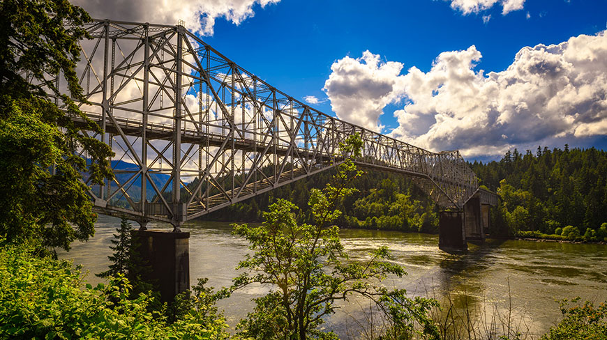 The steel truss Bridge of the Gods spans the Columbia River in Oregon, surrounded by lush green forests under a partly cloudy blue sky.
