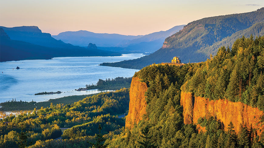 The sun sets over the Columbia River Gorge in Oregon, illuminating the Vista House perched on a cliffside overlooking the water.