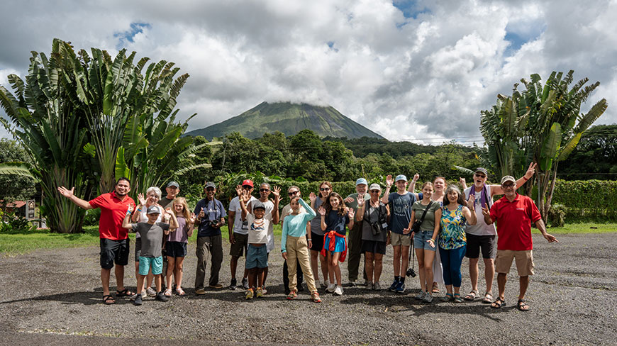 A group of travelers waves for a picture in front of a cloud-covered volcano and large traveler's palms in Costa Rica.