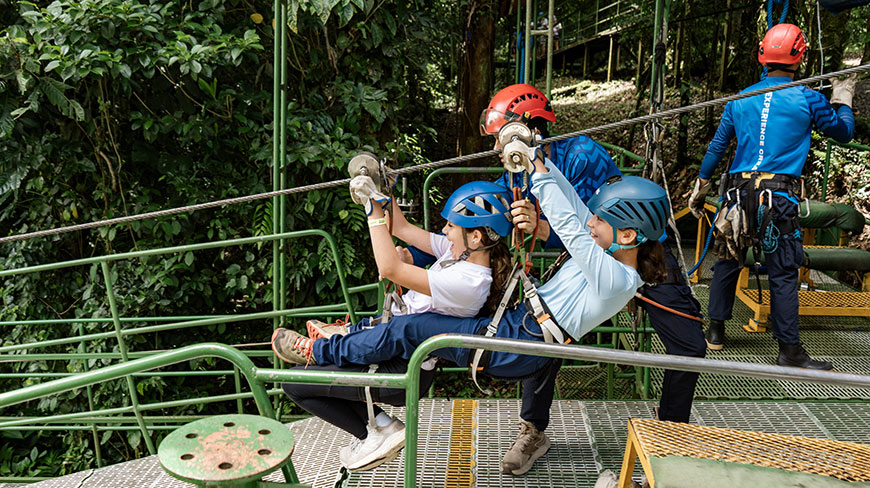 Two excited girls in helmets and harnesses sit on a platform, preparing to zipline with an instructor in Costa Rica.