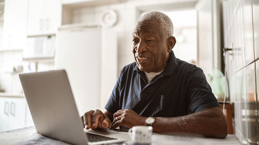 An older man smiles while participating in an online outdoor photography class on his laptop at a table.