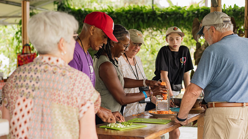 A group of travelers participates in a home cooking class in Costa Rica, grating carrots and chopping vegetables together at a wooden table.