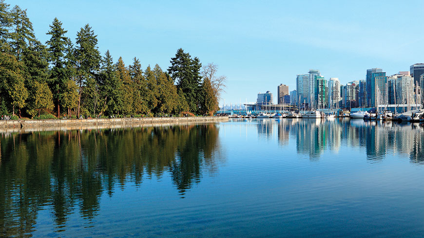 The Vancouver city skyline and forested Stanley Park are reflected in the calm harbor waters under a clear blue sky.