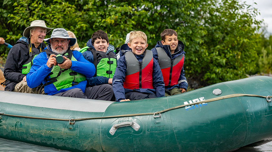 A multigenerational family smiles while enjoying a scenic float rafting tour in a large green inflatable raft in Alaska.