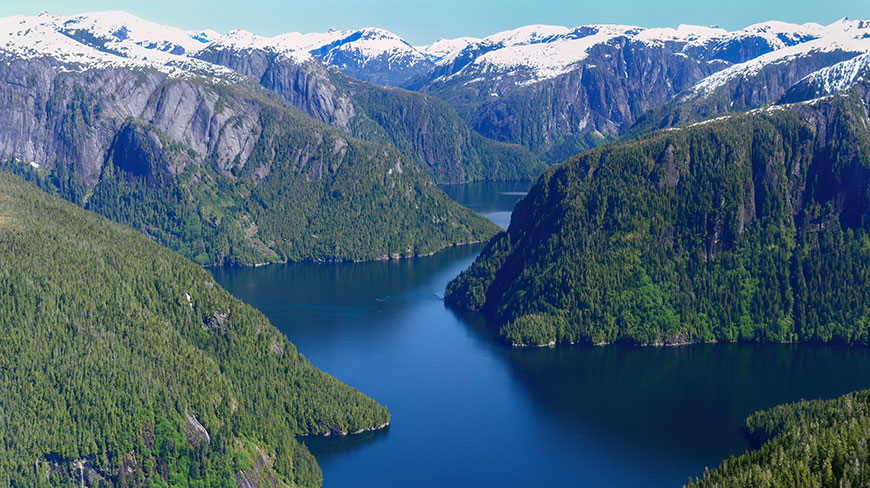 Sunlight shines on the snow-capped mountains and green forests surrounding a tranquil fjord in Misty Fjords National Monument, Alaska.