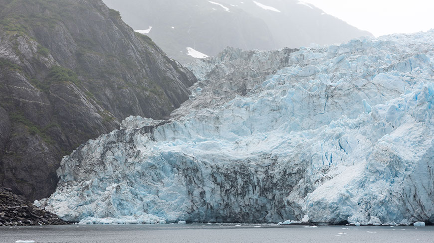 A massive glacier meets the water at the base of a steep, rocky mountain in Kenai Fjords, Alaska.