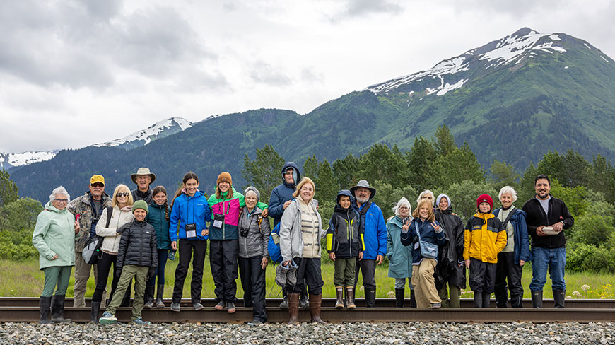 A group of travelers poses for a photo on railroad tracks in Alaska, with snow-capped mountains in the background.