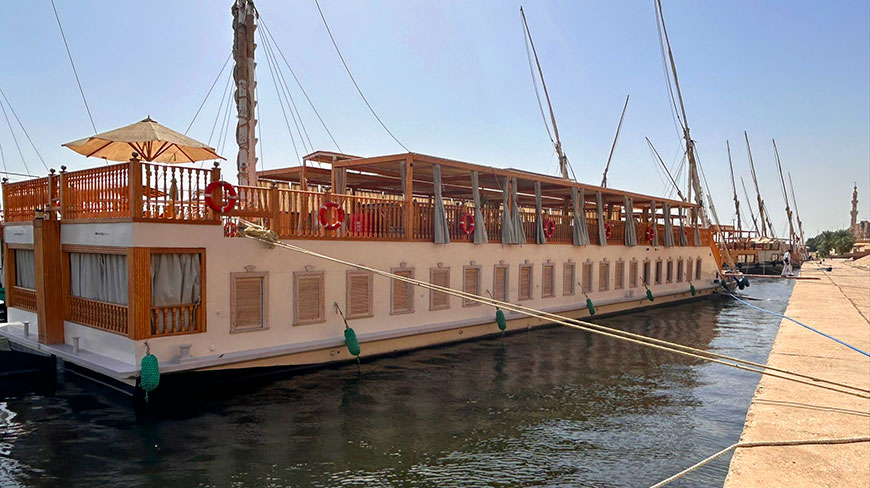 A modern dahabeya sailing vessel is moored at a stone dock along the Nile River in Egypt.