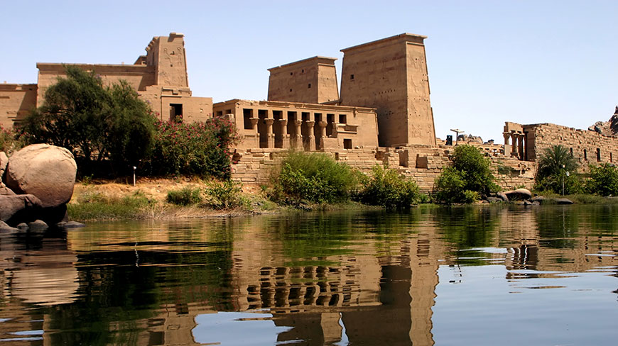 The ancient Temple of Philae in Egypt as seen from the Nile River, its stone structures reflected in the calm water.
