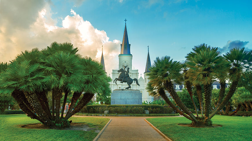 A walking path leads through a grassy park with palm trees toward the historic St. Louis Cathedral in New Orleans, Louisiana.