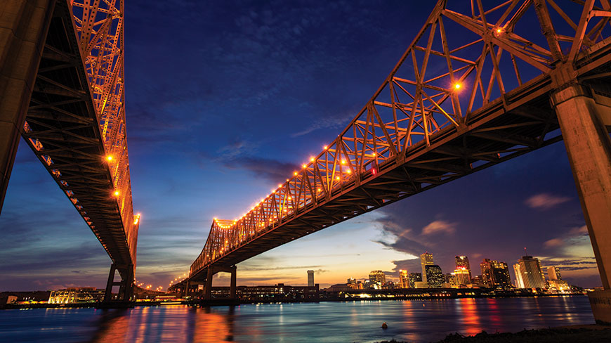 The Crescent City Connection bridge, illuminated with golden lights, spans the Mississippi River at dusk with the New Orleans skyline in the background.