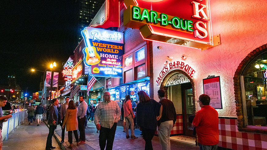 A vibrant nighttime scene on Beale Street in Memphis, with people walking past the bright neon signs of barbecue joints and bars.