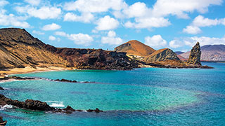 The iconic Pinnacle Rock juts out of a turquoise bay on the Galápagos Islands, with volcanic hills under a partly cloudy sky.