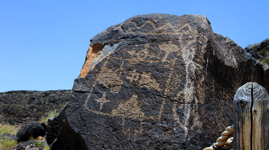 Ancient petroglyphs are carved into a large volcanic rock in New Mexico under a clear blue sky next to a wooden post.