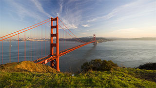 The Golden Gate Bridge in San Francisco is viewed from a nearby hillside, with the city skyline visible in the background.