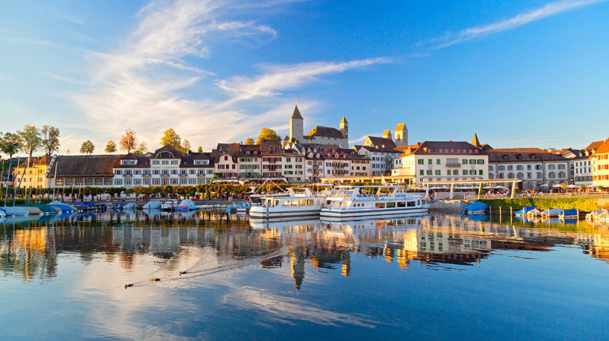 The waterfront of Rapperswil, Switzerland, with boats docked on the calm lake and historic buildings reflecting in the water under a blue sky.