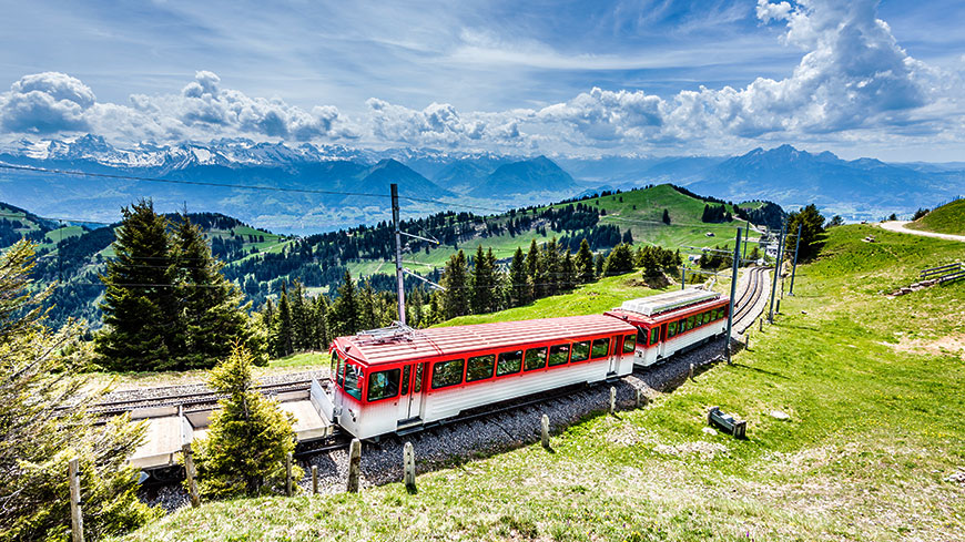 A red and white train travels up a lush green mountainside in Switzerland, with a panoramic view of the snow-capped Alps behind it.