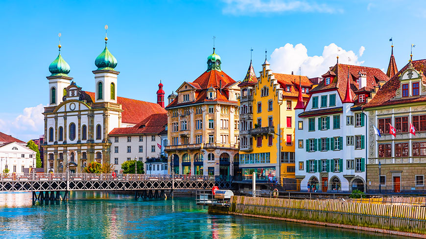 Colorful historic buildings and the Jesuit Church line the Reuss River in Lucerne, Switzerland, under a clear blue sky.