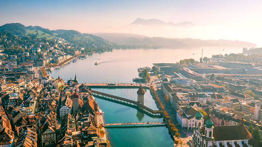 An aerial view of the city of Lucerne, Switzerland, with its iconic Chapel Bridge spanning the river against a backdrop of mountains.