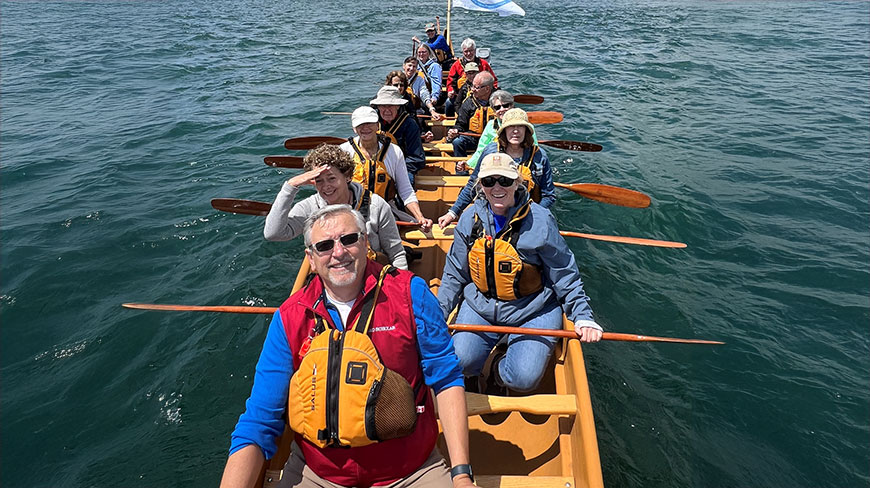 A group of people wearing life vests paddle a large canoe together on the water between Michigan and Ontario.