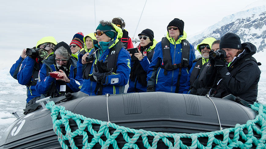 A group of tourists in winter gear on a zodiac boat take photos of the icy landscape from the water.