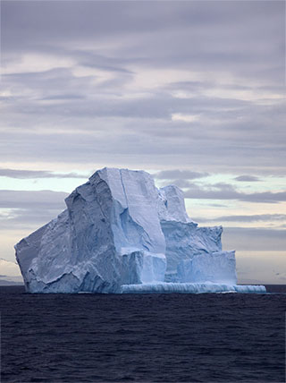 A large, craggy iceberg with blue shadows floats in the dark ocean water of the Drake Passage under a cloudy sky.