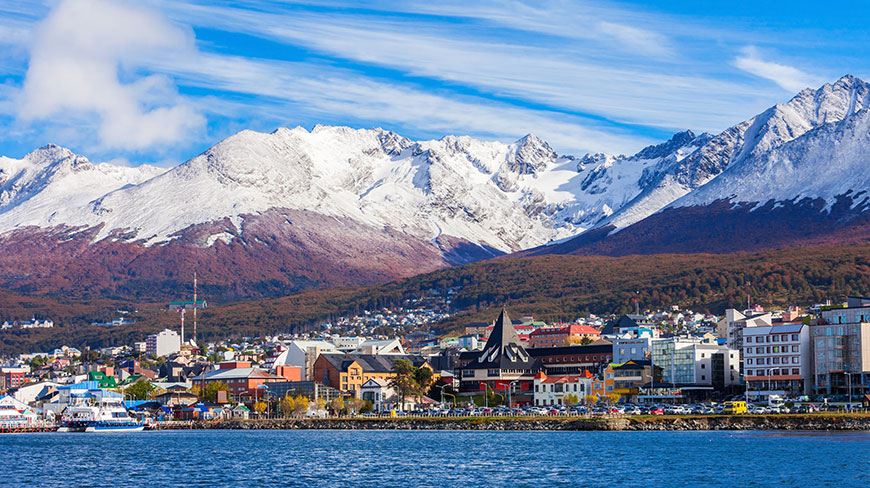 The colorful coastal town of Ushuaia is nestled at the base of a dramatic, snow-covered mountain range under a bright blue sky.