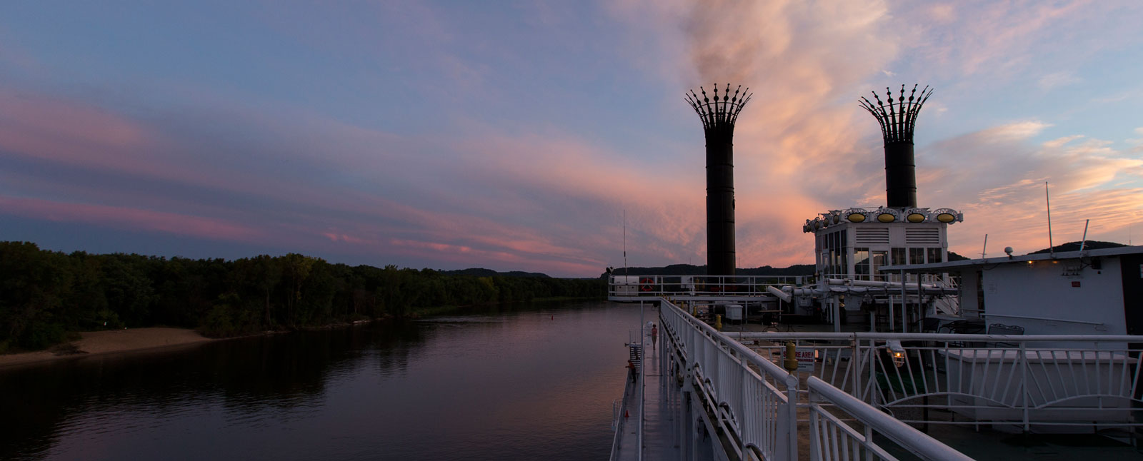 A view from the deck of a riverboat with decorative smokestacks cruising a river under a vibrant pink and blue sunset.