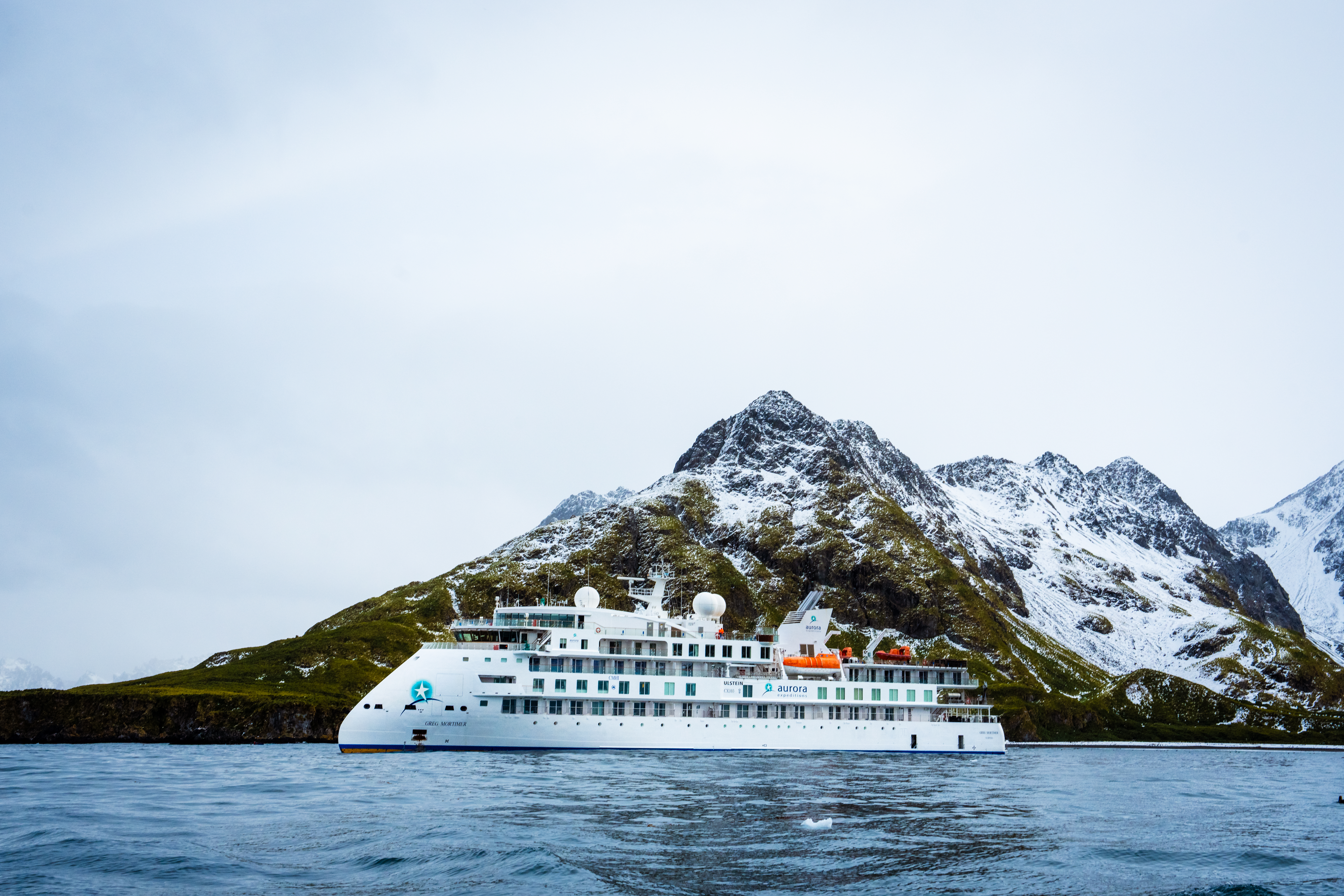 The Greg Mortimer expedition ship floats in the water in front of a large, snow-dusted mountain range under an overcast sky.