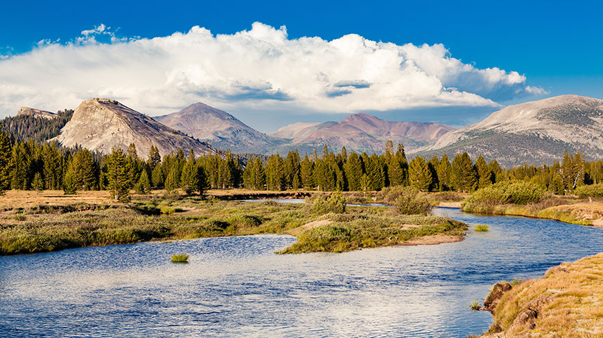 A river winds through Tuolumne Meadows in California's Yosemite National Park, with granite domes and mountains rising under a partly cloudy sky.