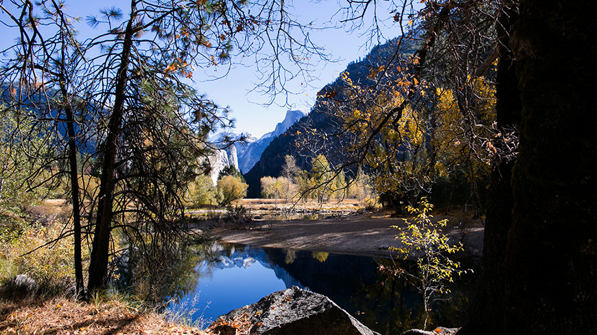 A tranquil river in Yosemite, California reflects granite domes and a distant waterfall, framed by pine and deciduous trees in autumn.