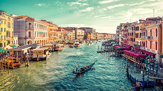Gondolas and boats navigate the Grand Canal in Venice, Italy, which is lined with colorful, historic buildings under a partly cloudy sky.
