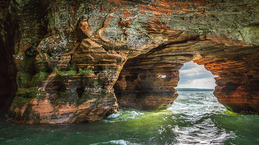 Choppy green water splashes against eroded red rock sea caves on Lake Superior in Wisconsin.