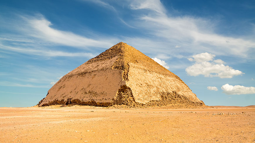 The Bent Pyramid at Dahshur in Egypt stands in the desert under a partly cloudy blue sky.