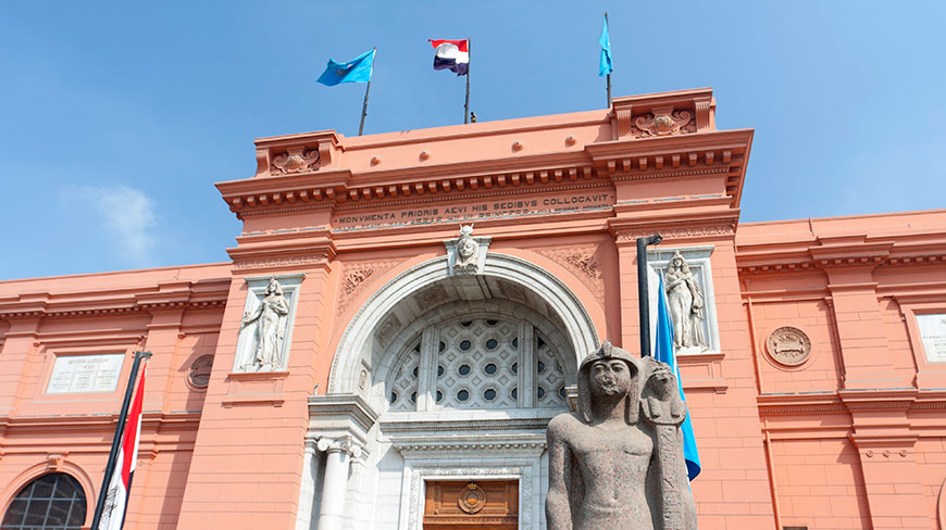 The ornate, salmon-colored facade of the Egyptian Museum in Egypt, with a large pharaoh statue guarding the grand arched entrance.