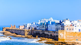 The white and blue buildings of the coastal city of Essaouira, Morocco, are seen from the water on a sunny day.