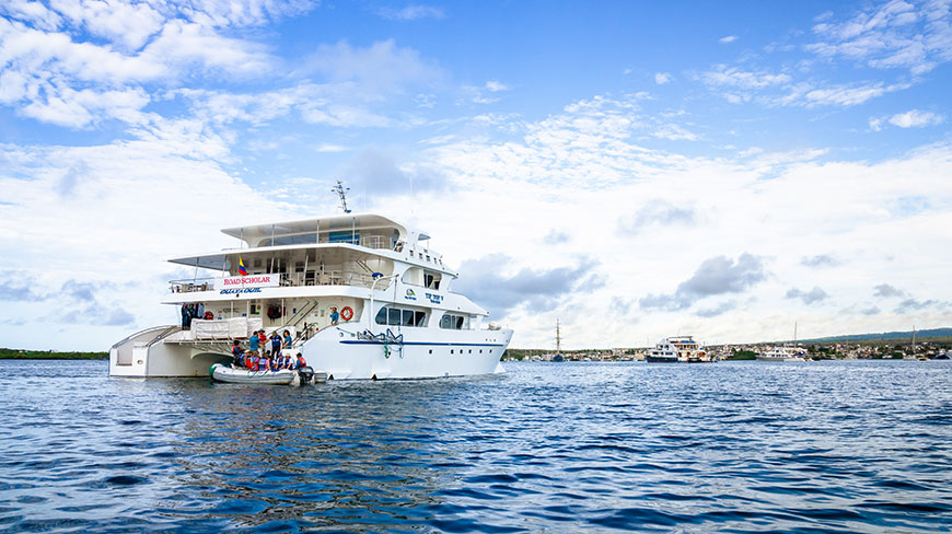 A white catamaran cruise ship floats on the water with a smaller boat alongside in the Galápagos Islands, Ecuador under a blue, cloudy sky.