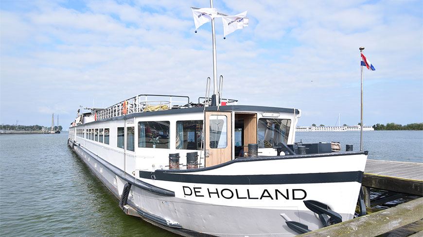 The De Holland ship is docked at a pier in the Netherlands on a partly cloudy day.