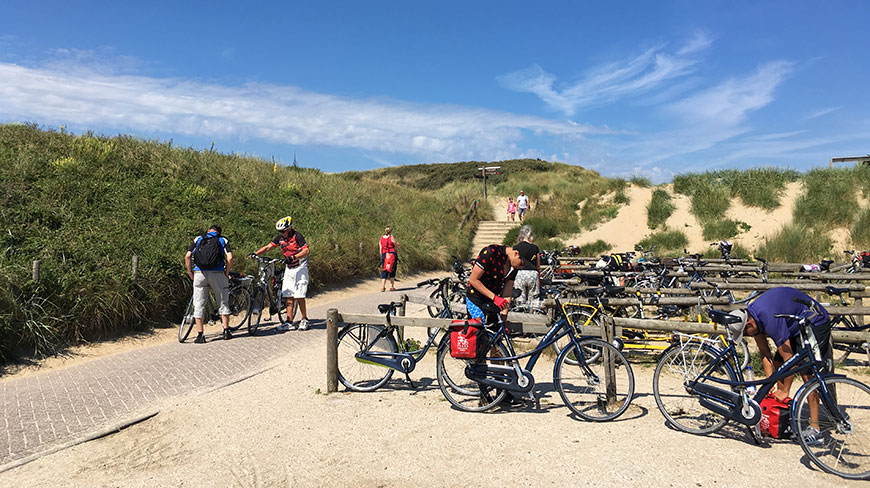 Cyclists park their bikes in a rack at the bottom of a sandy dune path in the Netherlands.