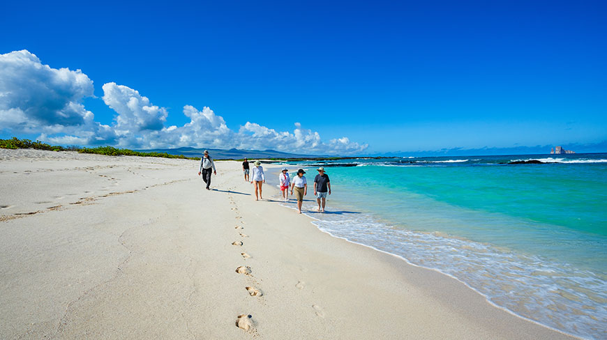 A group of travelers walks along a white sand beach beside turquoise water on Isla Lobos in the Galápagos Islands.