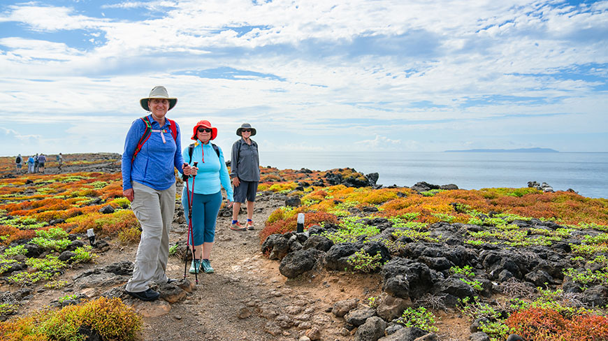 Three women in hiking gear smile while on a rocky coastal trail covered with colorful succulent plants in the Galápagos Islands, Ecuador.